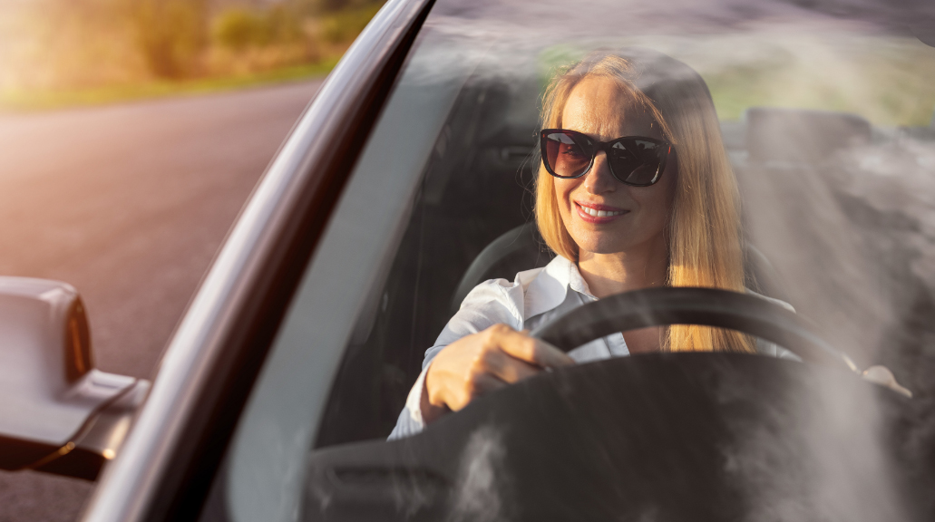 Woman in sunglasses and formal wear looking at side mirror while driving car. View through windshield. Sunny weather outdoors.