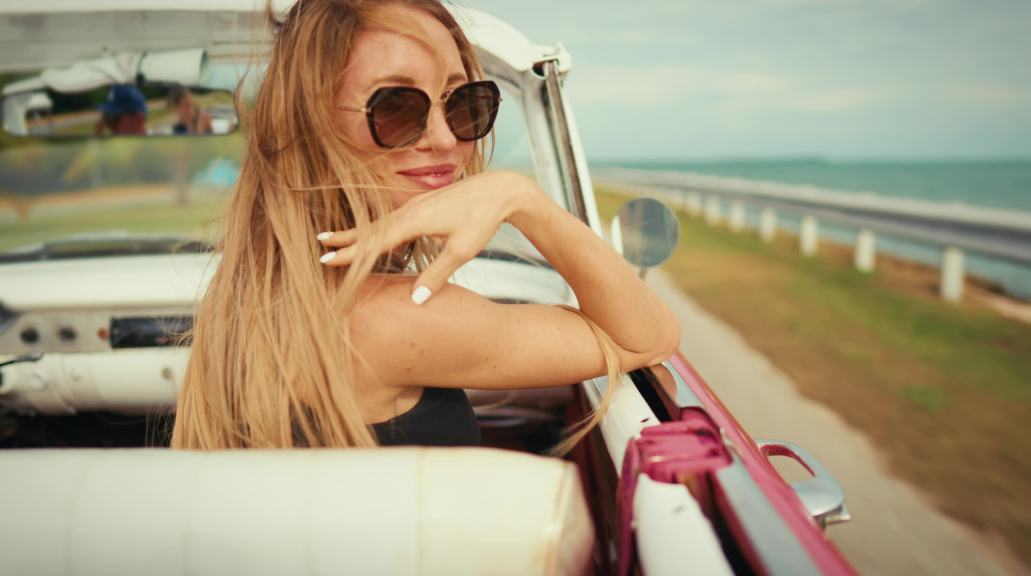 Happy blonde woman in cabriolet, long hair swaying by wind, joyful smile of face wearing sunglasses