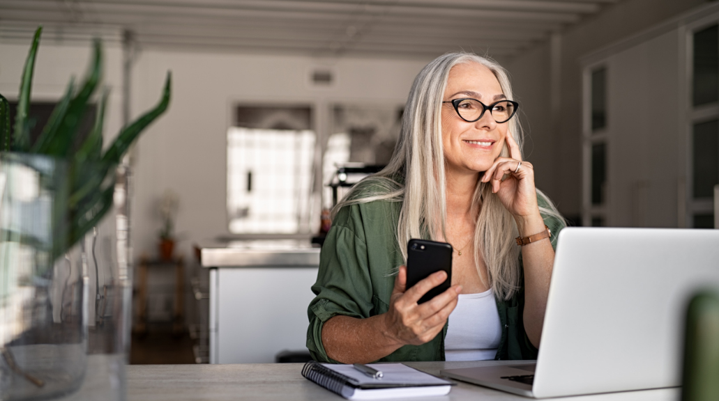 Mature woman smiling wearing glasses, holding her phone and sat with laptop