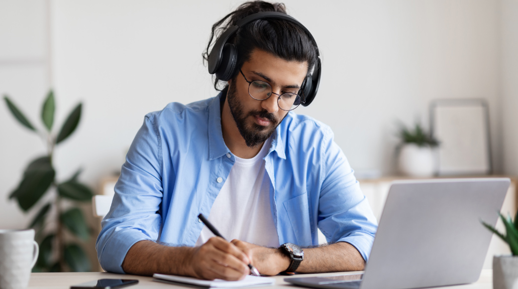 Man wearing glasses and headphones writing at home while using his laptop