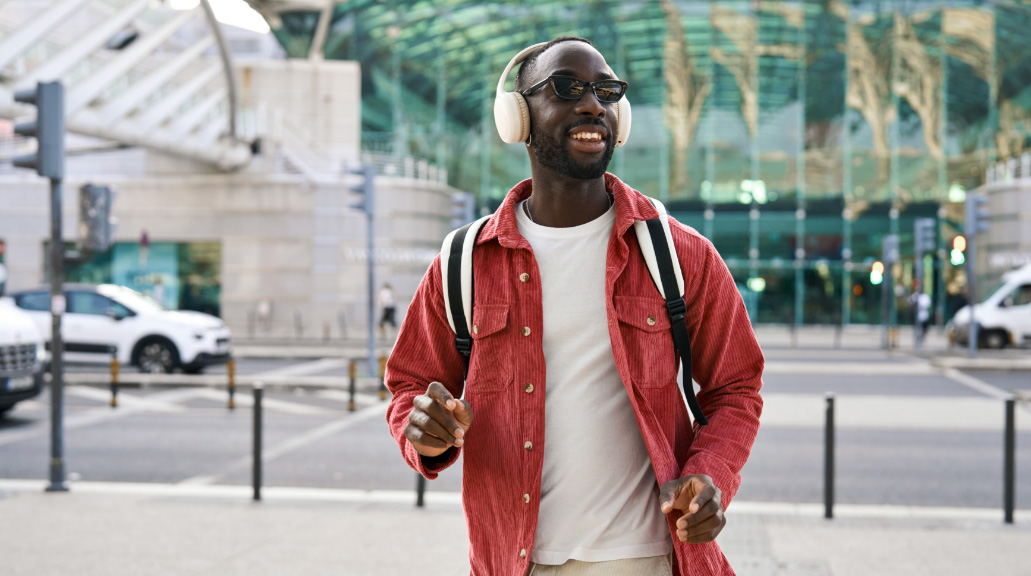 Happy cool stylish young man wearing headphones, sunglasses and backpack having fun listening to music and dancing walking on city street outdoors