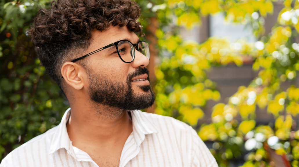 Portrait of a young man wearing glasses, smiling and looking away in a garden with a blurred background