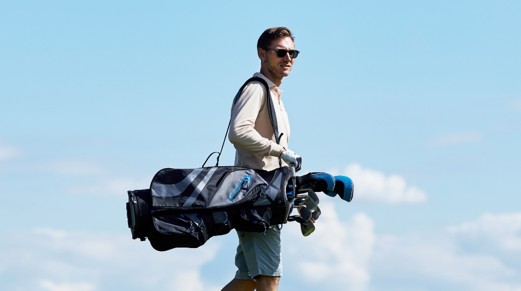 Side view portrait of sporty man carrying golf bag walking on green field against sky wearing sunglasses