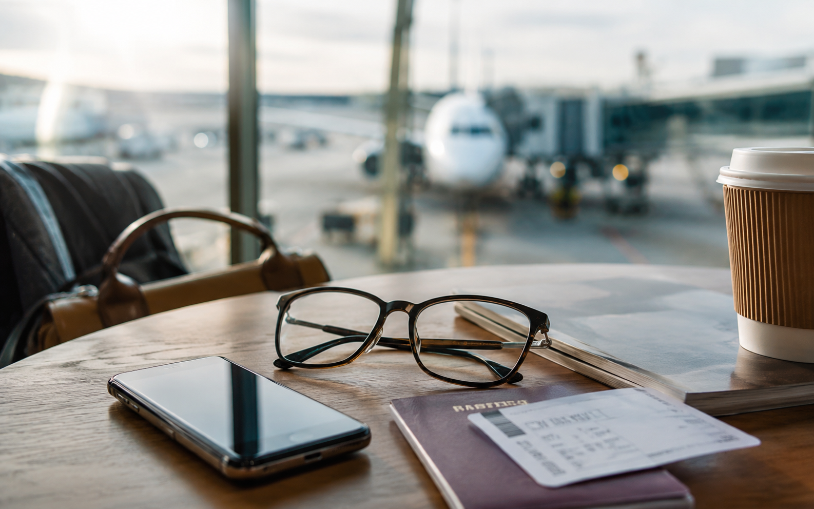Prescription glasses on an airport table beside a phone, passport, boarding pass, coffee cup, and book before a flight.
