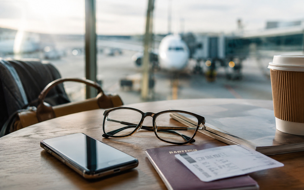 Prescription glasses on an airport table beside a phone, passport, boarding pass, coffee cup, and book before a flight.