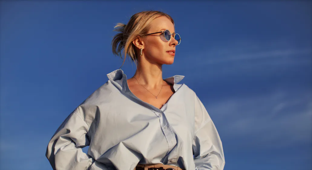 Modern woman in an oversized shirt and sunglasses stands outdoors against the sky