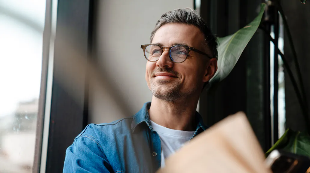 Portrait of adult smiling man in glasses looking out the window while sitting with book in cafe