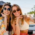 Stylish smiling women relaxing having fun at the beach wearing sunglasses, using smartphone to take selfie picture