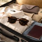 Sunglasses packed in an open suitcase with summer clothes, suncream, a book, passport, and glasses case for a holiday.